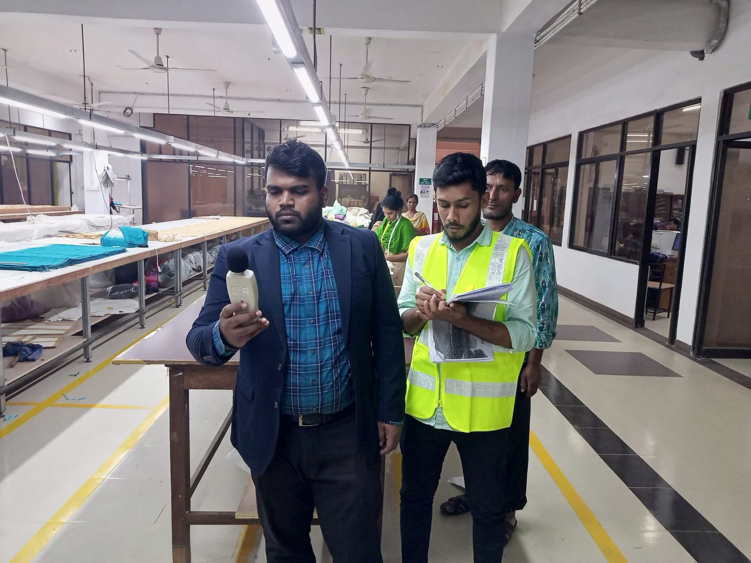 Two team members using a handheld sound level meter and recording field notes on a textile production floor